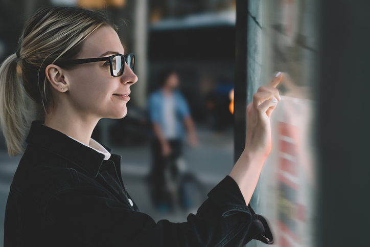 Smiling female standing at big display with advanced digital technology. Young woman touching with finger sensitive screen of interactive kiosk for find information while standing on street in evening.