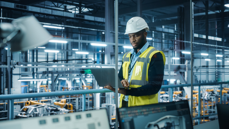 Car Factory: Professional Male Automotive Engineer Wearing Hard Hat, Walking, Using Laptop. Monitoring, Control, Equipment Production. Automated Robot Arm Assembly Line Manufacturing Electric Vehicles