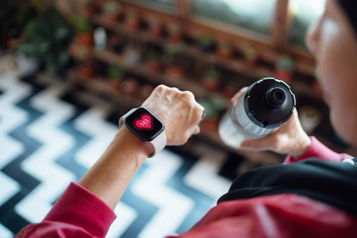 Over the shoulder view of senior Asian woman resting after exercising at home, looking at her smart watch, using fitness tracker app and measuring pulse. Maintaining healthy fitness habits. Elderly wellbeing, health, wellness and technology concept.