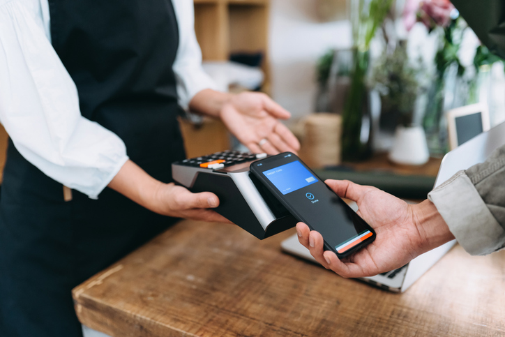 Close up of young Asian man shopping at the flower shop. He is paying with his smartphone, scan and pay a bill on a card machine making a quick and easy contactless payment. NFC technology, tap and go concept.